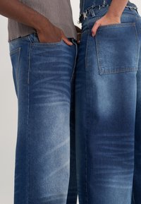 Dva páry modrých džín s gradientem od světlé po tmavou barvu, široké nohavice a standardní kapsy, zobrazené těsně vedle sebe.