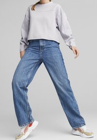 Felpa corta di colore viola chiaro con maniche lunghe, abbinata a jeans blu a vita alta e gamba larga, completata da sneaker bianche con dettagli rossi.