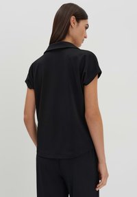 Zwarte blouse met korte mouwen, een kraag en gladde, lichtgewicht stof. De achterkant heeft een subtiele gebogen zoom en minimale naden.