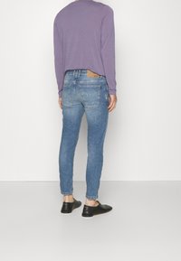 Jeans ajustados de mezclilla azul con un diseño desgastado, cinco bolsillos y un parche de cuero marrón en la parte trasera. Combinados con zapatos negros sin cordones.
