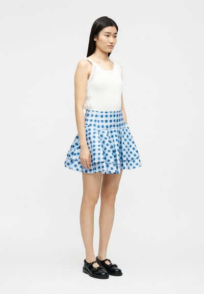 Γυναίκα όρθια, φορώντας λευκό αμάνικο τοπ με δαντέλα στα πλάγια, μπλε και λευκή καρό φούστα με βολάν και μαύρα παπούτσια Mary Jane.