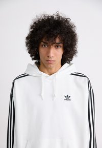 Moletom branco feito de mistura de algodão, apresentando um bolso canguru, riscas pretas nas mangas e um logótipo verde da Adidas no peito.