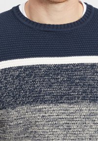 Neulepusero, jossa on teksturoitu tummansininen pohja ja vaakasuora valkoinen raita, joka siirtyy alaosassa vaaleampaan harmaaseen.