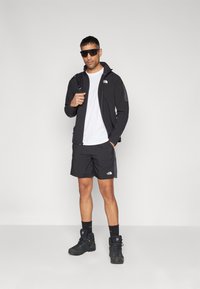 Chaqueta atlética negra con cremallera, camiseta blanca y pantalones cortos negros. Lleva puesto botas negras y gafas de sol. Logo notable en la chaqueta y los pantalones cortos.