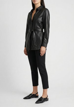 Femme portant une chemise en cuir noire ceinturée, un pantalon noir slim et des chaussures slingback noires pointues, debout devant un fond uni.