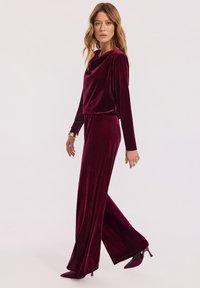 Kobieta idąca w głębokoczerwonym aksamitnym topie z długimi rękawami i szerokich spodniach, w dopasowanych czubkowych szpilkach oraz złotej bransoletce.