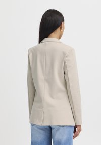 Beige přizpůsobený blazer s jedním zadním rozparkem, límečkovým designem a dlouhými rukávy, vystavený s džíny se světle modrou praním.