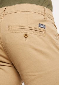 Pantalon en coton beige avec une poche arrière et un bouton, arborant une étiquette « BLEND ». Le tissu a une texture lisse et un design simple.