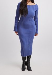 Donna che indossa un abito midi a maglia blu a maniche lunghe e righe, abbinato a stivali neri con tacco, su uno sfondo neutro.