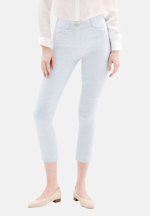 Femme portant un jean skinny court bleu clair, un chemisier blanc et des chaussures beiges à petits talons, debout les mains détendues le long du corps.