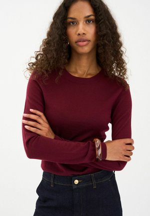 Pulover burgundy cu mâneci lungi, decolteu rotund, textura tricotată, asortat cu blugi închisi la culoare. Modelul poartă o brățară maro la încheietura mâinii.