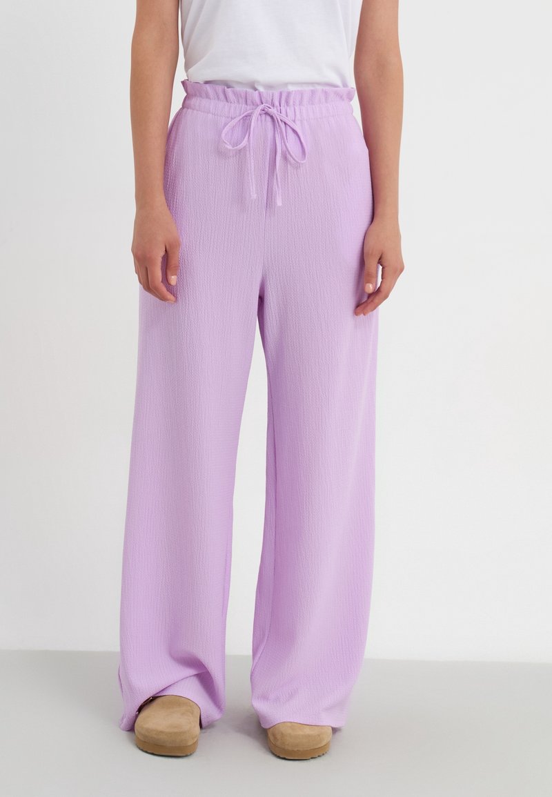 Pantalons larges violet clair en tissu texturé, taille élastique avec cordon de serrage et coupe décontractée. Portés avec des sabots beiges.