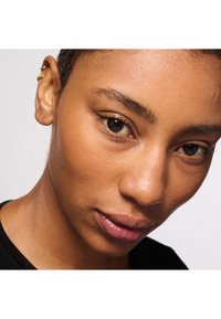 Zbliżenie na twarz z gładką skórą. Osoba ma krótkie, ciemne włosy i nosi czarną koszulę. Charakterystyczne szczegóły to złote kolczyki.