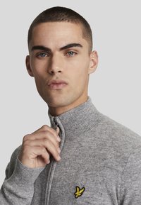 Maglione grigio con zip e colletto a coste, caratterizzato da un piccolo logo giallo ricamato sul petto. Realizzato in un morbido tessuto testurizzato.