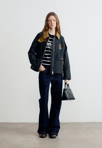 Γυναίκα που φοράει μαύρο σακάκι με καφέ γιακά, ριγέ πουκάμισο, σκούρα τζιν με φαρδιά πόδια, κρατώντας μαύρη τσάντα, στέκεται μπροστά σε απλό φόντο.