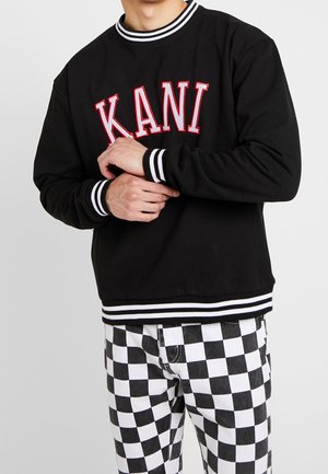 Sweat-shirt noir avec le logo "KANI" en rouge, poignets et ourlet rayés. Porté avec un pantalon à carreaux noir et blanc. Finition en tissu lisse.