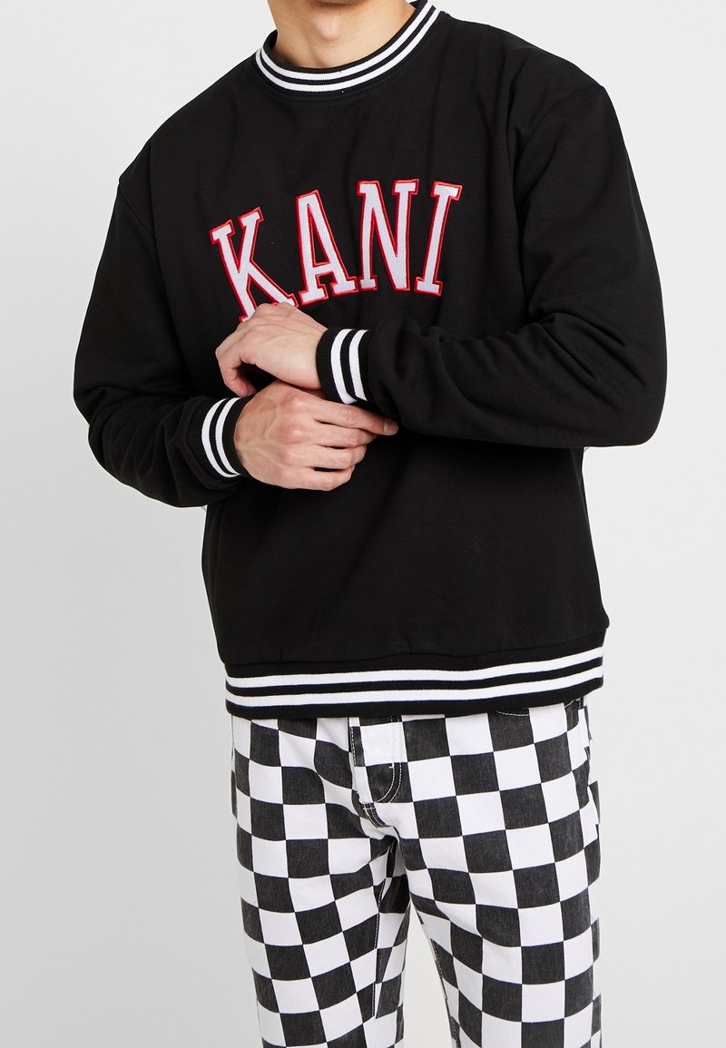 Felpa nera con logo "KANI" in rosso, polsini e orlo a strisce. Indossata con pantaloni a quadri bianchi e neri. Finitura del tessuto liscia.