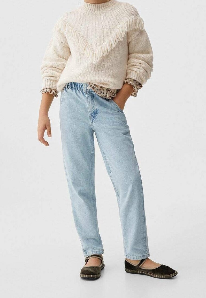 Personne portant un pull en maille crème par-dessus un chemisier fleuri, un jean bleu clair et des chaussures noires style Mary Jane, debout avec une main dans la poche.