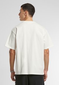 Tricou alb din bumbac, cu croi lejer, mâneci scurte și guler rotund. Partea din spate are o textură netedă și detalii minime.