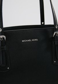 Zwarte leren tote bag met een gestructureerde afwerking, zilveren hardware, voorzien van dubbele handvatten en het geperste "Michael Kors" logo aan de voorkant.