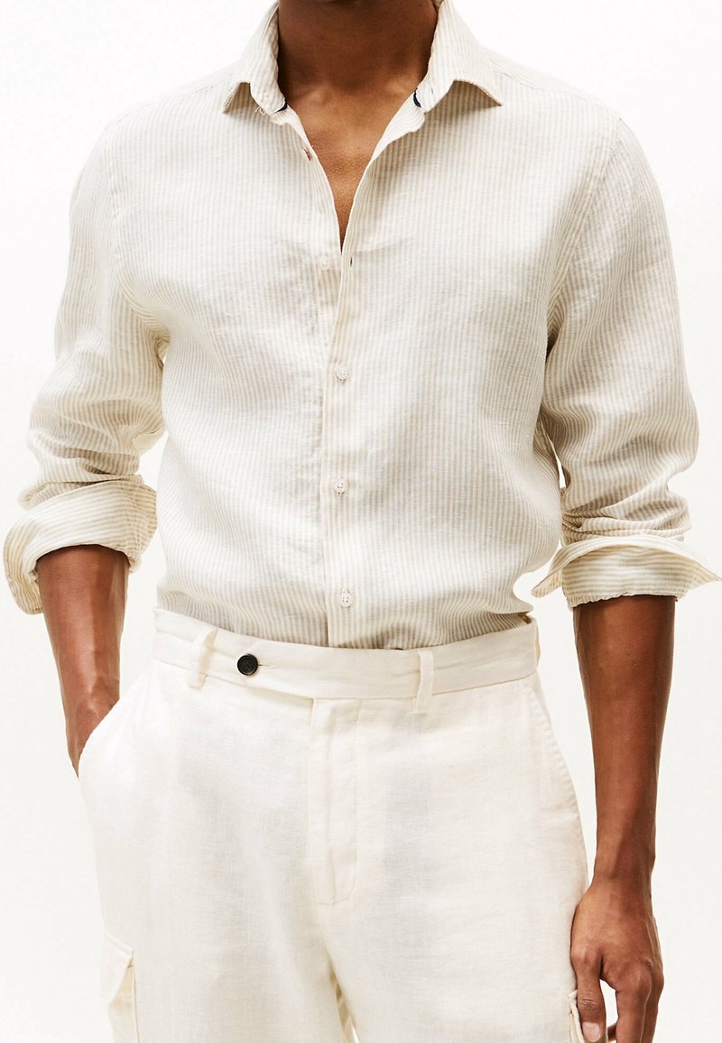 Beige gestreiftes Leinenhemd mit langen Ärmeln, Knopfverschluss vorne und hochgeklappten Manschetten. Kombiniert mit hellbeigen Hosen mit einem schwarzen Knopf.