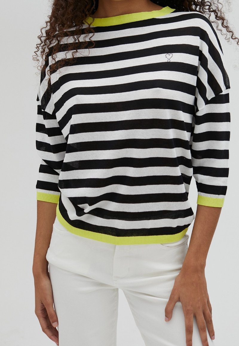 Gestreifter Pullover in Schwarz und Weiß mit lockerer Passform, featuring einem leuchtend grünen Kragen und Saum sowie einem kleinen dekorativen Herzelement.