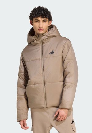 Junger Mann trägt eine beige Steppjacke mit Kapuze und passende beige Hose mit Logo, steht vor einem schlichten hellen Hintergrund.