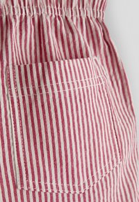 Lähivõte punase ja valge vertikaalselt triibulise kangast, millel on õmmeldud tasku ja kummiköitega kogutud vöökoht.