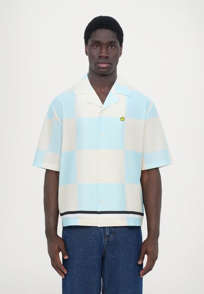 Camisa de botones de manga corta en un patrón de cuadros celeste y crema, con un pequeño parche de sonrisa amarillo y una franja negra en el dobladillo.