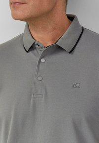 Grijze polo met een gestructureerde stof, twee knopen en een zwarte rand bij de kraag. Heeft een klein logo aan de linkerkant.