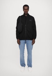Giacca nera con zip e maniche a costine, abbinata a jeans azzurri chiari e sneakers bianche. Borsa a tracolla nera di piccole dimensioni sulla spalla.