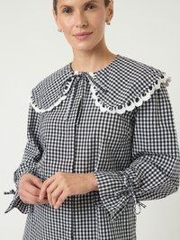 Svart og hvit gingham-bluse med stor hvit blondekrage og samlet ermer, med en knytedetalj ved halsen og håndleddene.