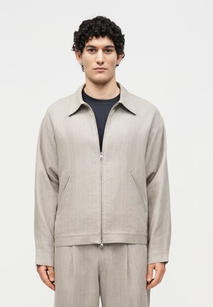 Mand med krøllet hår iført beige lynlåstrøje og matchende bukser, står mod en ensfarvet hvid baggrund og vender fremad.