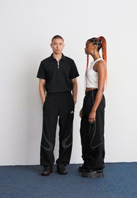 Svart glidelås polo og svarte Adidas joggebukser med hvite detaljer båret av en person; den andre personen i hvit crop topp og svarte bukser med teksturert design.