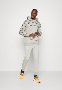 Sudadera deportiva gris con patrón de logo negro, pantalones de chándal grises a juego y zapatillas de correr amarillas brillantes. Sosteniendo una botella de agua.