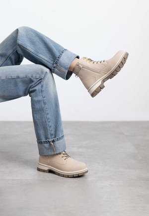 Botas de tobillo beige con cordones y suelas de goma, con ojales dorados y acentos marrones en contraste; usadas con jeans de mezclilla azul arremangados.