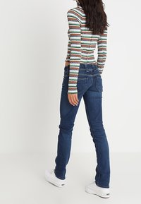 Blå denimjeans med smal passform, sidofickor och diskret bakstygn, tillsammans med en långärmad topp med flerfärgade ränder och vita sneakers.