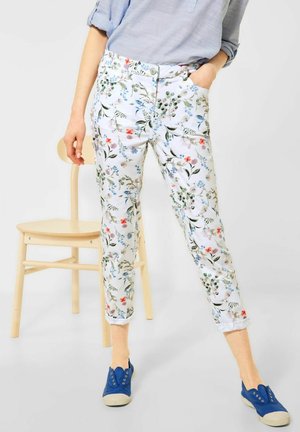 Personne portant un pantalon court blanc à fleurs, une chemise bleu clair aux manches retroussées et des chaussures en toile bleu à enfiler, debout à côté d'une chaise en bois.