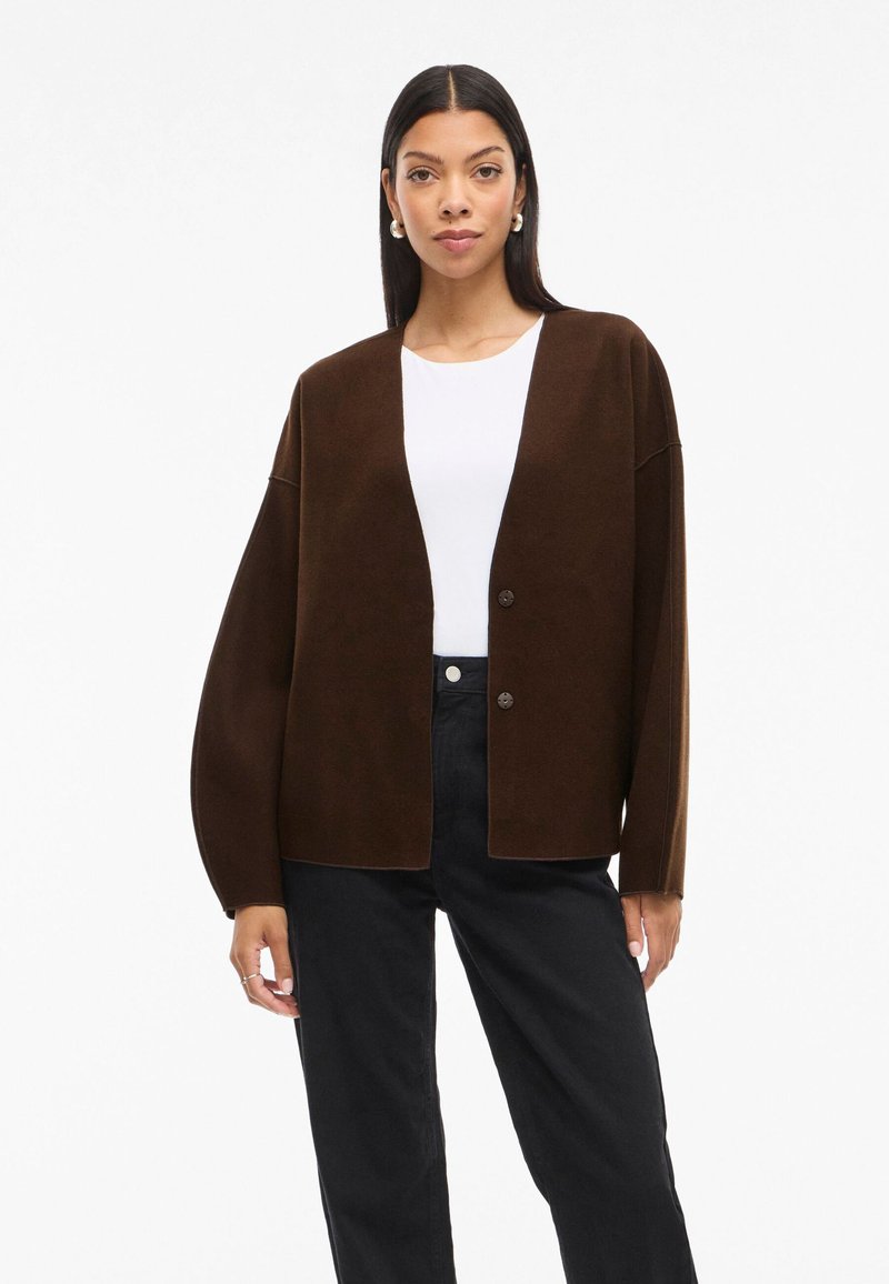 Rudas perdėtas megztinis su plačiomis rankovėmis, dviem mygtukais priekyje, dėvimas virš baltos marškinių ir juodų kelnių. Lygi tekstūra, minimalistinis dizainas.