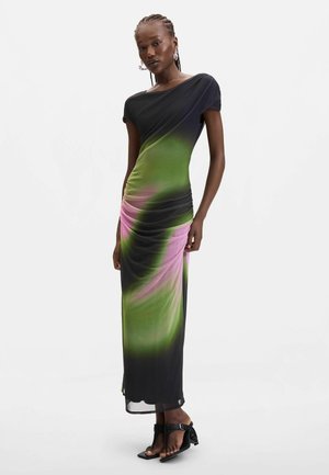 Femme debout dans une robe ajustée à manches courtes, longueur cheville, avec un motif dégradé vert, rose et noir, portant des sandales noires.