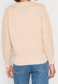 Persona che indossa un maglione a maglia testurizzata beige con motivi a diamante e jeans di denim blu, mostrata di spalle.