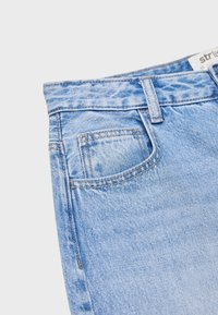 Jean en denim bleu clair en gros plan montrant la ceinture, la boucle de ceinture et la petite poche avant avec des coutures visibles sur un fond uni.