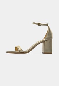 Χρυσό μεταλλικό πέδιλο με ψηλό τακούνι, με έναν ιμάντα πάνω στα δάχτυλα, ιμάντα αστραγάλου και ενιαίο τακούνι. Ομαλή υφή και μίνιμαλ σχεδιασμός.