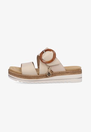 Sandales beige clair avec un design à large sangle, présentant un accent circulaire brun et un petit charm décoratif, reposant sur une semelle texturée.