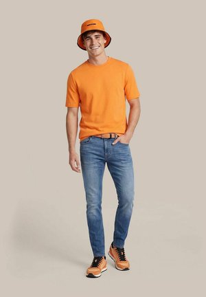 Man glimlacht, draagt een oranje vissershoed, oranje t-shirt, blauwe spijkerbroek, oranje sneakers, staat voor een effen achtergrond.
