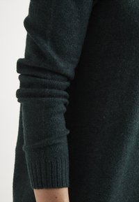 Ärm av en mörkgrön stickad tröja med ribbad manschett, något skrumpen ovanför handleden på en ljus hudton arm.