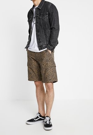 Hombre con una chaqueta de mezclilla negra, camiseta gráfica blanca, pantalones cortos cargo de leopardo y zapatillas negras de corte bajo con cordones blancos.