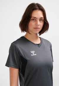 Mörkgrå sporttröja med korta ärmar, V-ringning, med en svart sidopanel och en vit logotyp på bröstet. Slät textur, lättviktigt tyg.