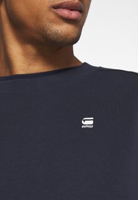 Mörkblå bomulls-T-shirt med rund hals, med en vit "RAW"-logotyp på bröstet, ribbad struktur och korta ärmar.