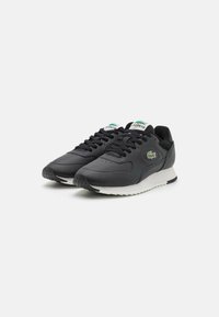 Chaussures de sport noires en matériau synthétique avec une finition lisse, dotées d'une semelle blanche, de lacets noirs et d'un détail de logo vert sur le côté.